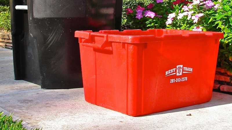 A red plastic recycling bin sitting on a concrete surface next to a black trash can, with pink flowers and green shrubs in the background. The red bin is branded with the "Best Trash" logo and phone number 281-313-2378.