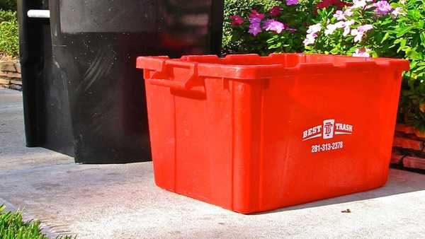 A red plastic recycling bin sitting on a concrete surface next to a black trash can, with pink flowers and green shrubs in the background. The red bin is branded with the "Best Trash" logo and phone number 281-313-2378.
